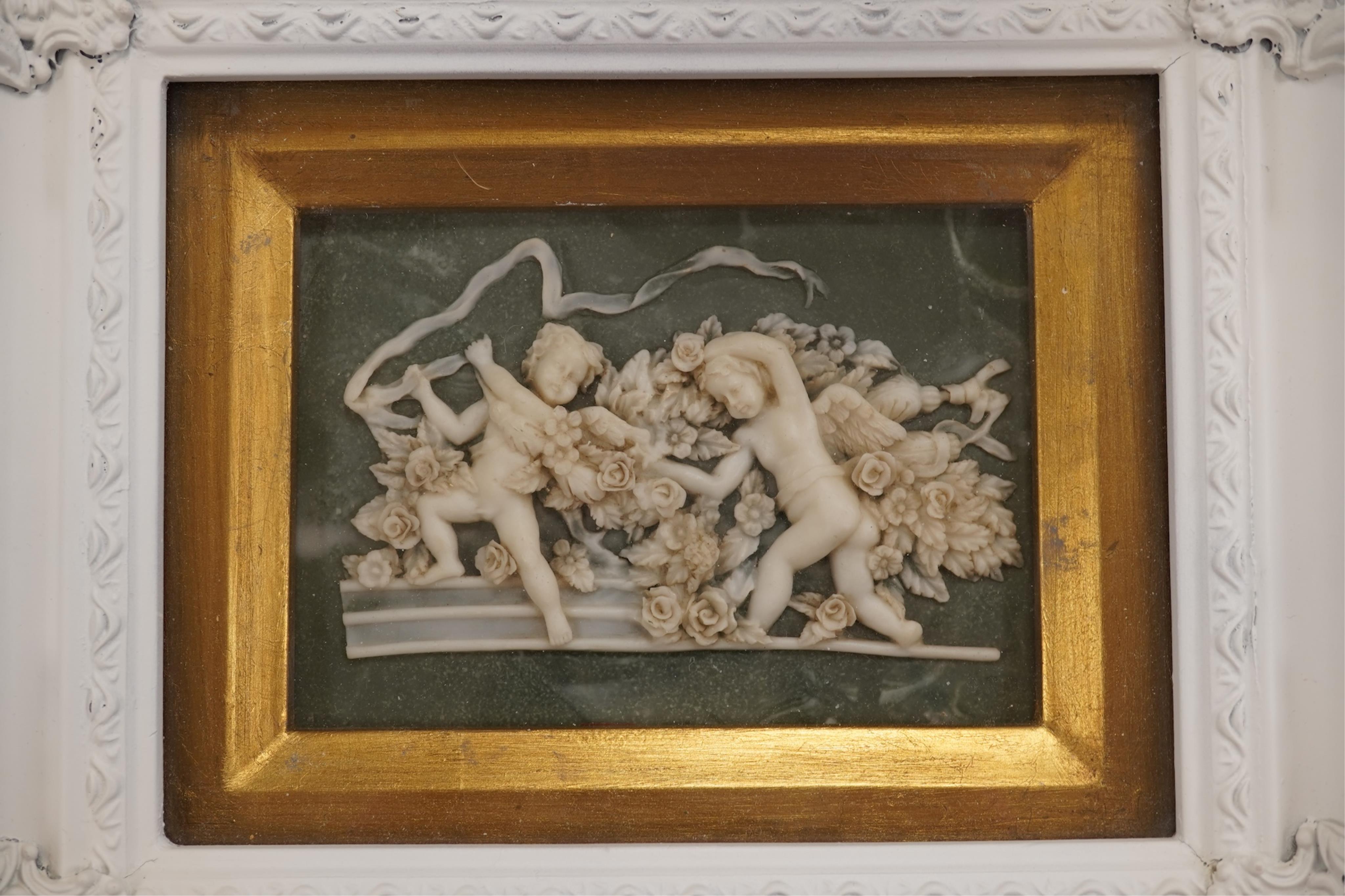 A pair of modern framed carved wax plaques of putti, housed in decorative painted frames, 27 x 32cm